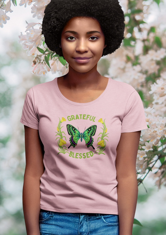 woman standing near tree wearing pink tee which reads grateful blessed, green butterfly in middle, yellow flowers on side of butterfly, available in many colors.